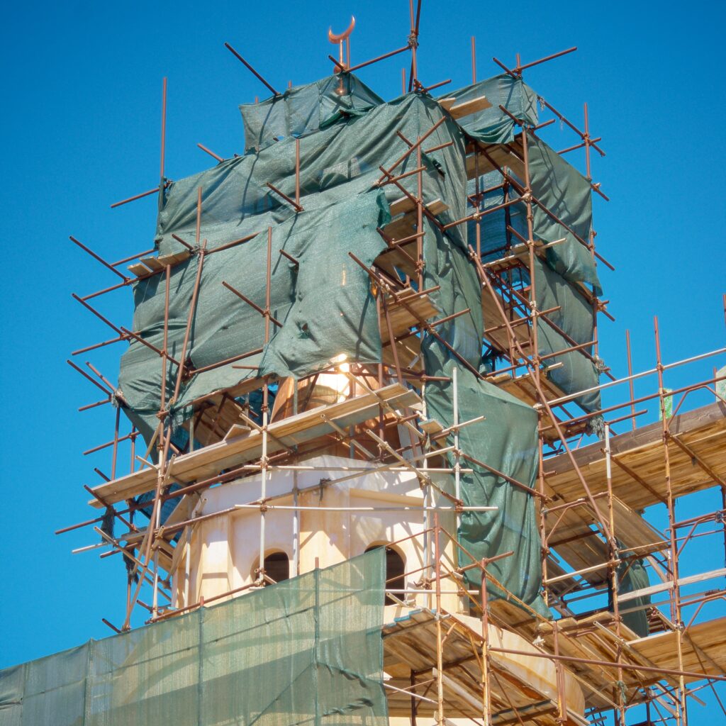 A tower under construction, covered with scaffolding and green tarps against a clear blue sky.