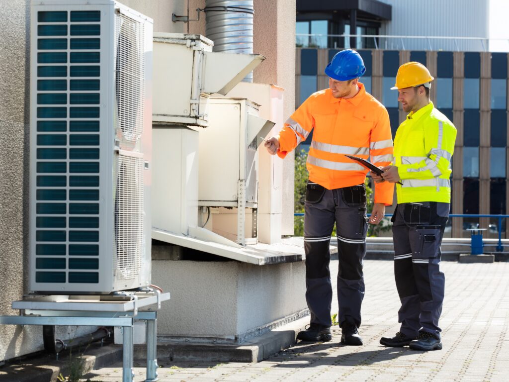 Two workers in safety gear inspect HVAC equipment on a rooftop.