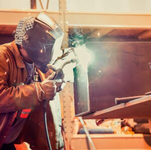 A welder in protective gear works on a metal piece, creating sparks and bright light during the welding process.