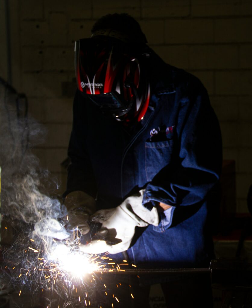 A person in protective gear is welding, producing sparks and smoke in a dimly lit workshop.